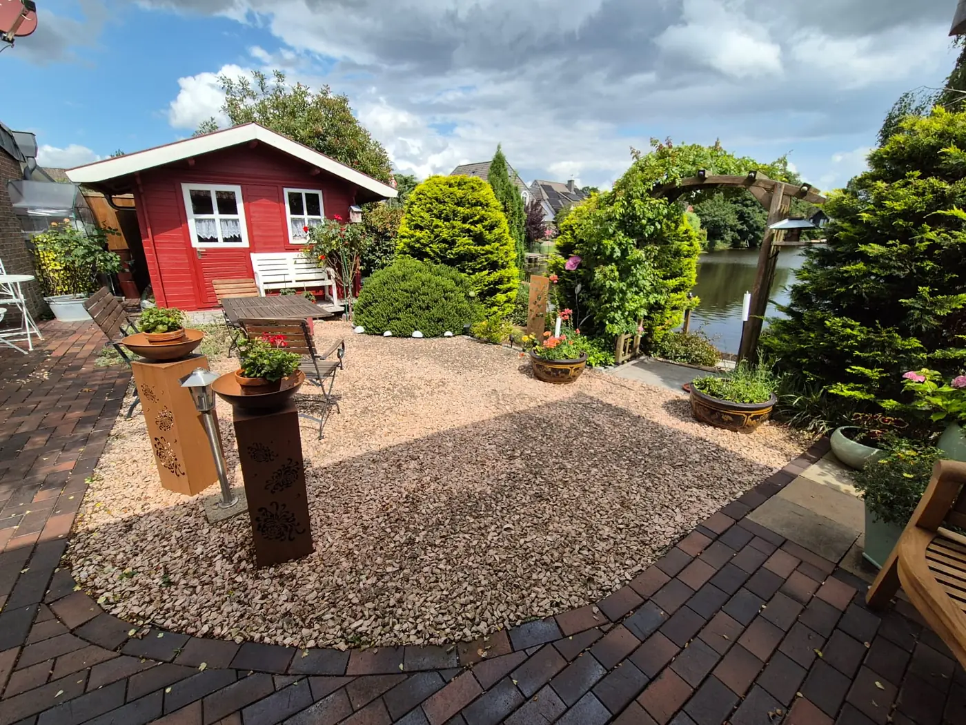 Gartenanlage mit Kiesfläche, Klinkerpflaster und Rost-Stelen vor einem Gartenhaus